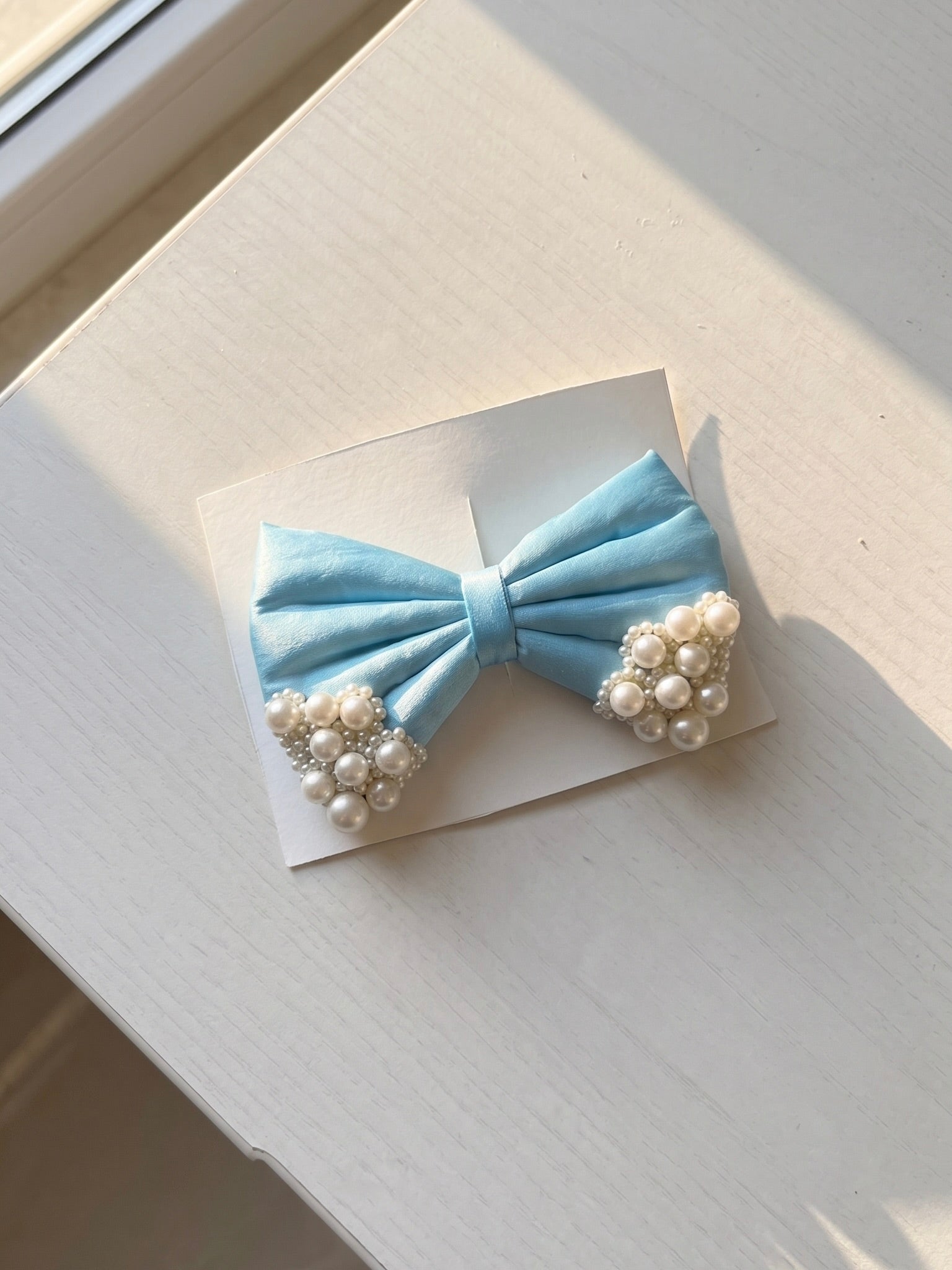 A light blue satin bow hair accessory with pearl clusters on both ends, displayed on a small white card on a light-colored wooden surface.